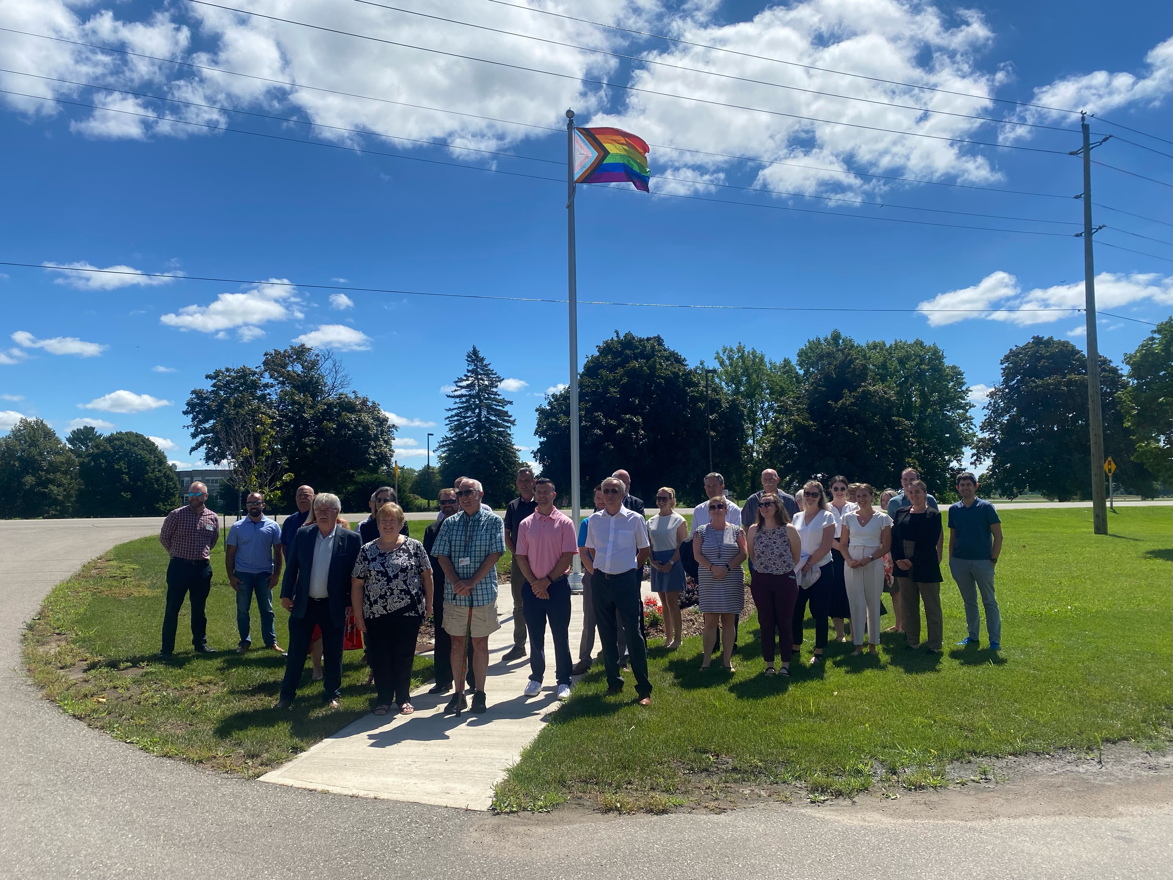 A group of people standing in front of a flag pole flying the Pride flag, there are clouds in the sky but the day is sunny. 